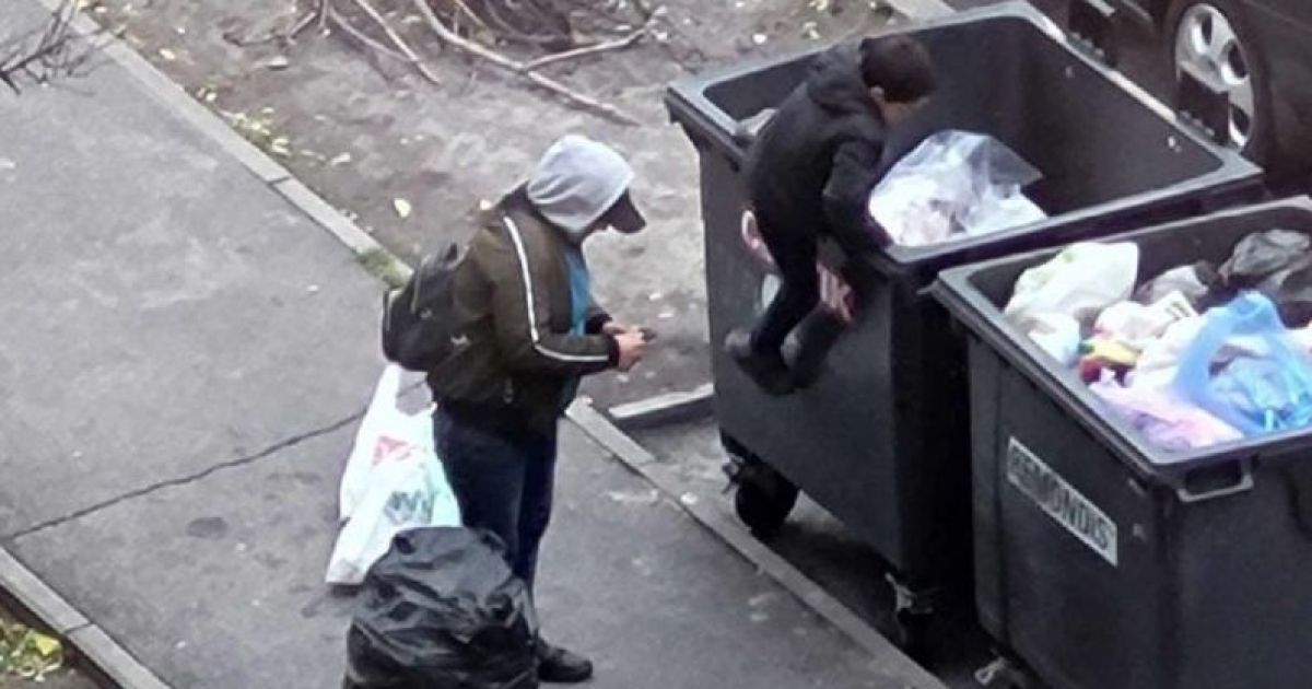 Мать подсаживает мальчика в мусорный бак.