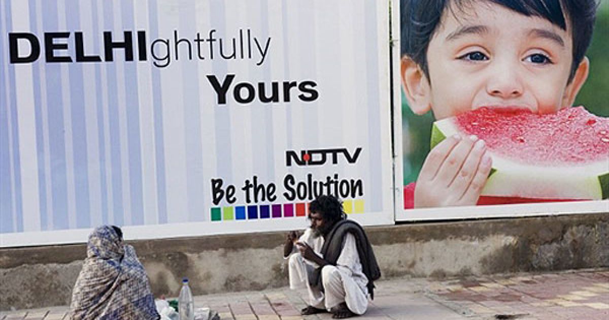 Індія, Нью-Делі. Безпритульна пара обідає на землі перед рекламним плакатом у Нью-Делі. В індійській столиці з 3 по 14 жовтня мають відбутися Ігри Співдружності, найбільша спортивна подія у місті з 1982 року. / © AFP