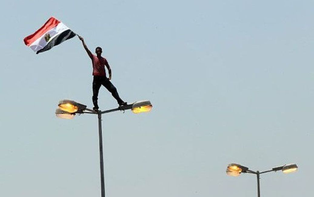 Єгипет, Каїр. Єгипетський демонстрант розмахує національним прапором, стоячи на вуличному ліхтарі на площі Тахрір в центрі Каїра. Десятки тисяч людей вийшли на площу Тахрір, щоб взяти участь в акції протесту. / © AFP
