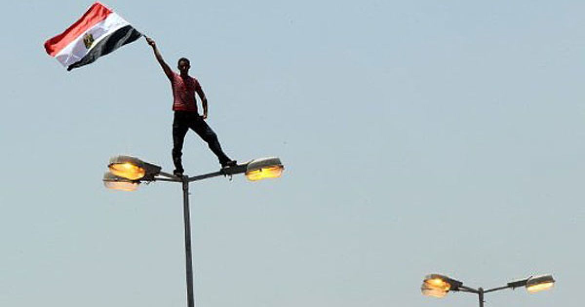 Єгипет, Каїр. Єгипетський демонстрант розмахує національним прапором, стоячи на вуличному ліхтарі на площі Тахрір в центрі Каїра. Десятки тисяч людей вийшли на площу Тахрір, щоб взяти участь в акції протесту. / © AFP