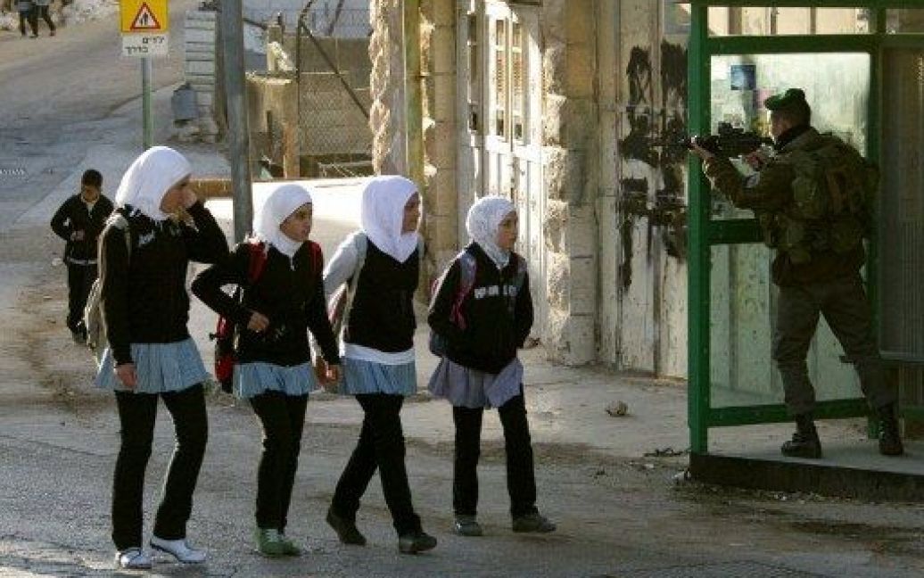 Хеврон. Палестинські дівчата проходять повз ізраїльського прикордонника, який патрулює вулицю біля палестинського будинку в центрі Хеврона на окупованому Західному березі. / © AFP