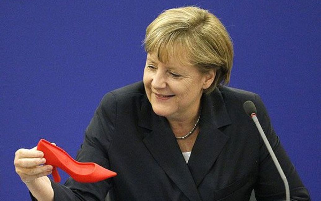 Німеччина, Берлін. Федеральний канцлер Німеччини Ангела Меркель роздивляється дверну підпору у вигляді червоного черевичка під час зустрічі з Асоціацією профспілок Німеччини. Черевичок подарували Меркель, як символ того, що сучасні жінки увійшли до професійного сучасного світу. / © AFP