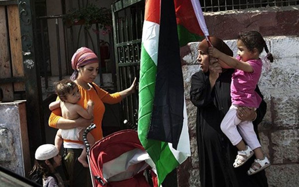 Єрусалим. Палестинська жінка (праворуч), чий будинок був окупований ізраїльськими поселенцями, несе на руках свою дитину, яка тримає палестинський прапор. Поруч із ними проходить ізраїльська жінка-поселенець, яка тепер мешкає у цьому спірному будинку у східному районі Єрусалиму Шейх-Джаррах. В Єрусалимі провели акцію протесту з нагоди першої річниці масового виселення палестинських родин з їхніх будинків. / © AFP
