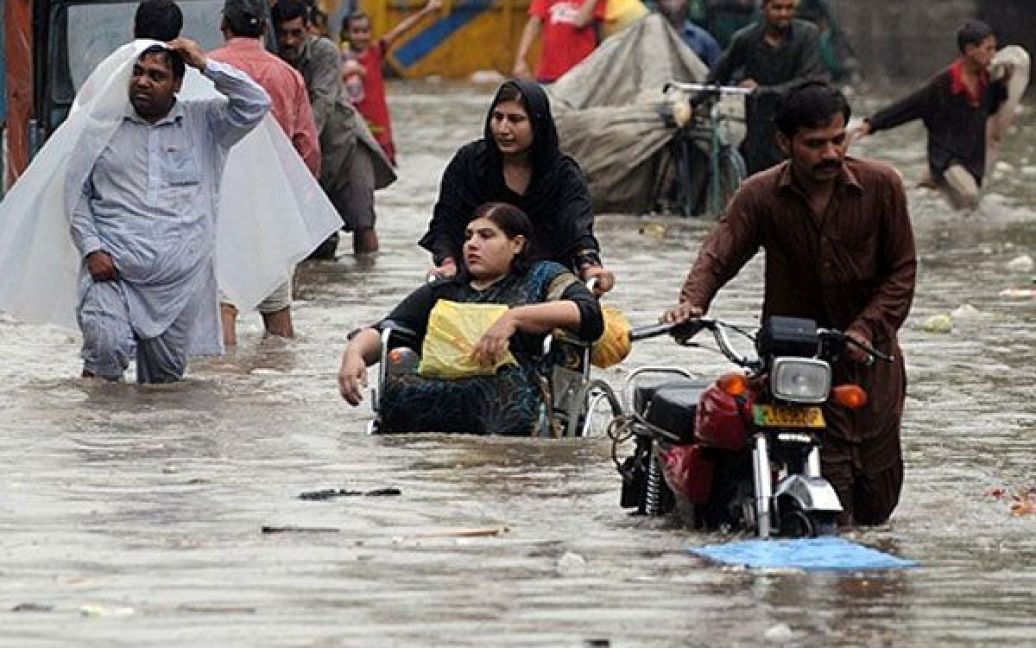 Пакистан, Лахор. Пакистанська жінка, яка штовхає інвалідне крісло зі своєю дочкою, та інші перехожі йдуть затопленою вулицею в Лахорі. Метеорологічне управління Пакистану прогнозує ще більш дощів протягом наступних двох днів. / © AFP