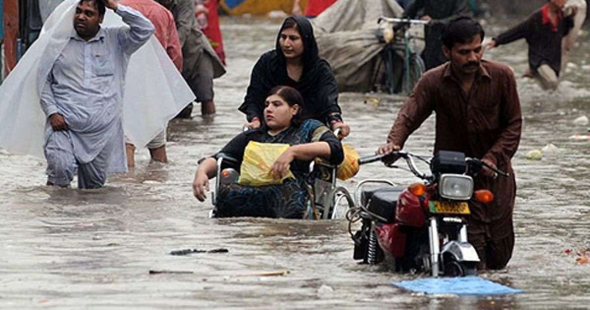 Пакистан, Лахор. Пакистанська жінка, яка штовхає інвалідне крісло зі своєю дочкою, та інші перехожі йдуть затопленою вулицею в Лахорі. Метеорологічне управління Пакистану прогнозує ще більш дощів протягом наступних двох днів. / © AFP