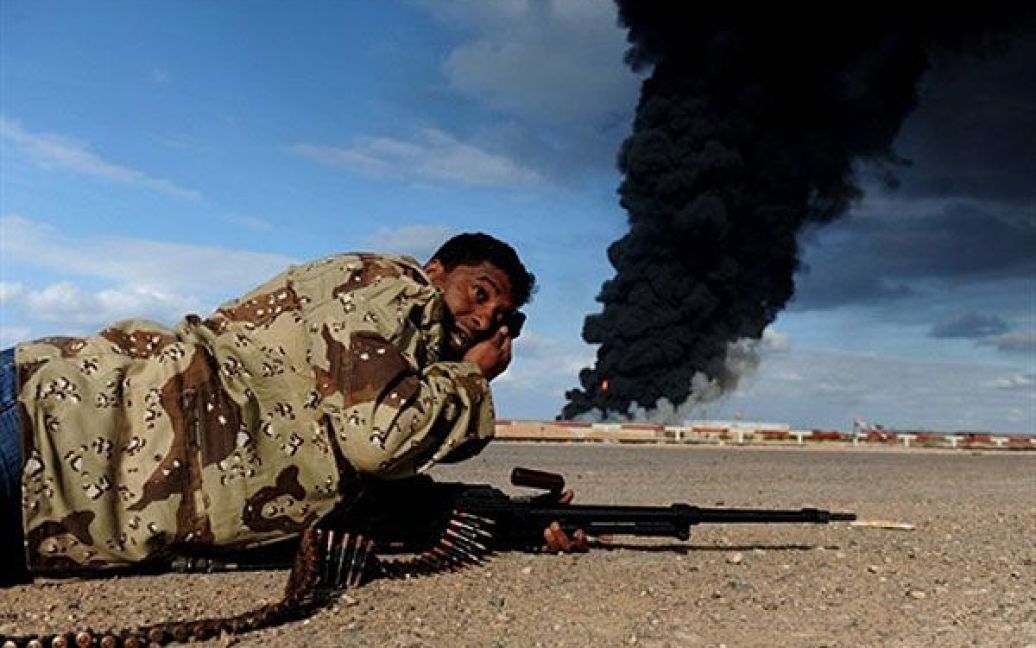 Лівійська Арабська Джамахірія, Рас Лануф. Лівійський бойовик-повстанець лежить на землі, доки снаряди урядових військ Муаммара Каддафі вибухають навколо, а чорний дим піднімається з пошкодженого нафтопроводу. / © AFP