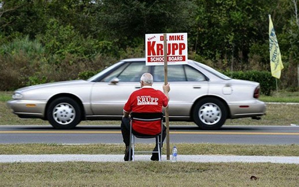 США, Тітусвілле. Доброволець тримає агітаційний плакат, сидячи поруч із виборчою дільницею у місті Тітусвілле, штат Флорида. У США почалось голосування на проміжних виборах. / © AFP