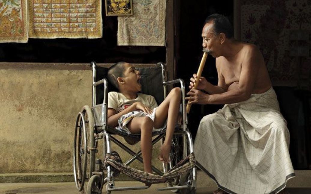 Музика любові. Фотографія зроблена у селі Тенганан, острів Балі. Чоловік грає на бамбуковій сопілці, щоб розважити дитину-інваліда, який не є його сином, але якого він любить як власного сина. Фото Аріо Вібісоно. / © National Geographic