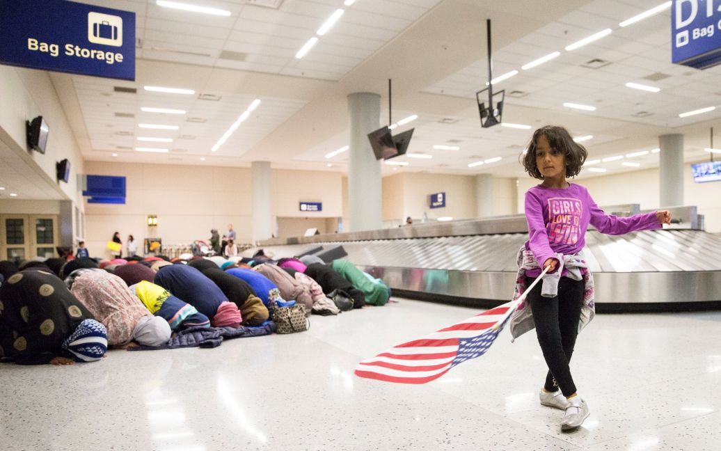 Молода дівчина танцює з американським прапором в зоні отримання багажу, в той час як жінки моляться позаду неї під час акції протесту проти антиімміграційного указу Дональда Трампа, в міжнародному аеропорту у Далласі. / © Reuters