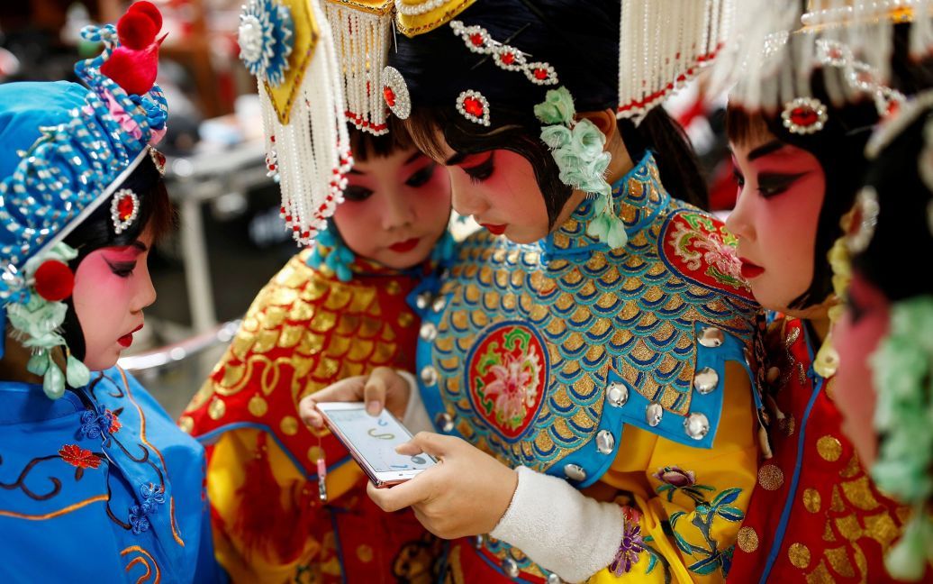 Учасниця групи артистів традиційної китайської опери грає в гру на своєму телефоні, а інші спостерігають за нею, під час антракту у Національній академії китайського театрального мистецтва в Пекіні, Китай. / © Reuters