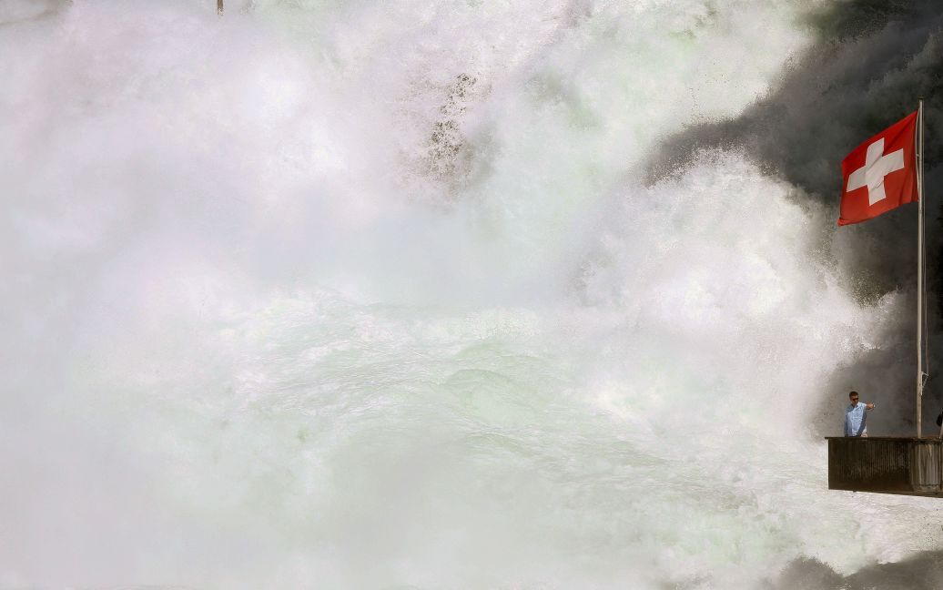 Национальный флаг Швейцарии висит на платформе рядом с Рейнским водопадом в то время, как человек делает на фоне его селфи. Рейнский водопад является крупнейшим водопадом в Европе. / © Reuters