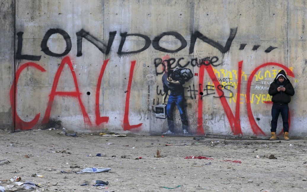 Мигрант разговаривает по телефону в лагере, известном как "Джунгли", в городе Кале на севере Франции. На стене вместе со знаменитым граффити Бэнкси, изображающего Стива Джобса-мигранта, написано по-английски: "Лондон зовет". Мигранты в Кале всеми способами пытаются попасть в Великобританию. / © Reuters