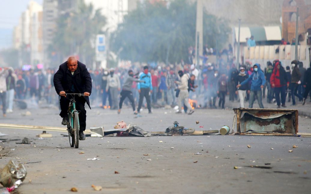 Людина їде на велосипеді в той час як протестувальники кидають каміння в бік поліції під час акції протесту поблизу місцевої адміністрації в місті Кассеріна, Туніс. Кілька тисяч молодих людей вийшли на протести після того, як тиждень тому молодик покінчив життя самогубством через те, що йому було відмовлено у громадській роботі. / © Reuters