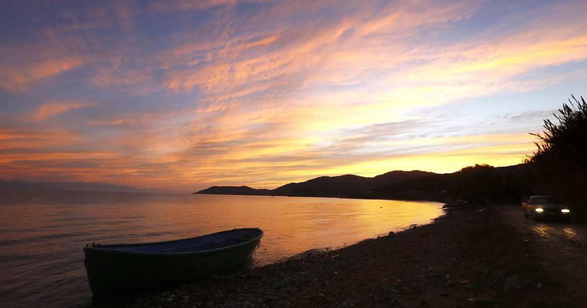 Місцевий фермер їздить на пікапі по дорозі у той час, як на пляжі стоїть дерев'яний човен біженців, на якому вони прибули до грецького острова Лесбос. Із 660 000 мігрантів, які потрапили до Греції в цьому році, більше половини прибули на Лесбос. В цьому році понад 3460 біженців загинули при перетині Середземного моря, 360 з них - в останні чотири місяці, 250 з них - в грецьких територіальних водах. / © Reuters