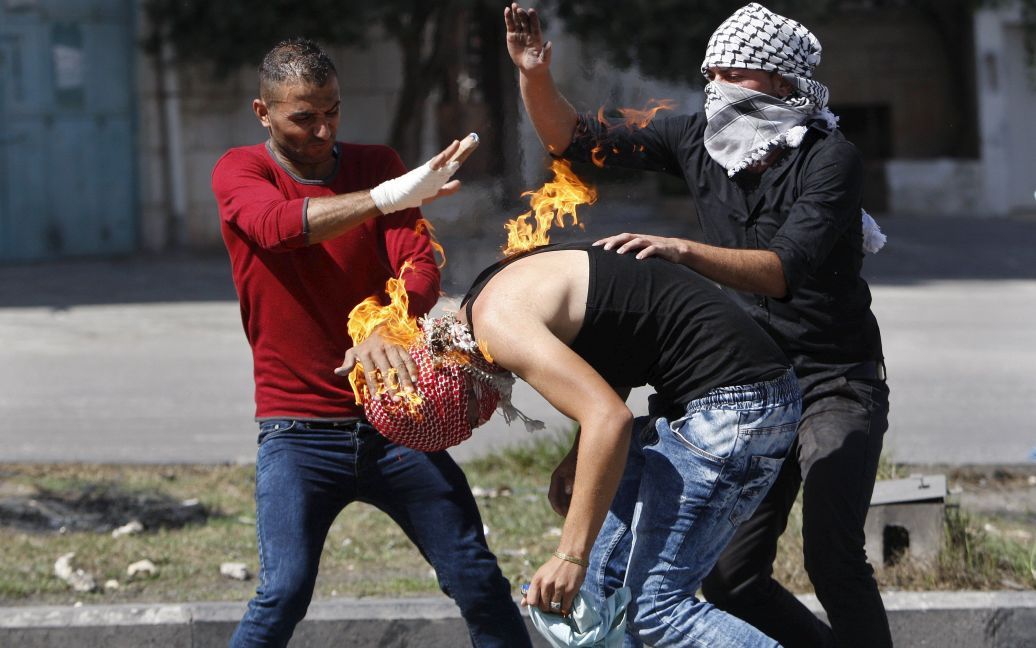 Палестинские демонстранты тушат огонь, пылающий на соотечественнике. Огонь вызвал коктейль Молотова, который демонстрант пытался бросить в израильских войск в ходе столкновений в городе Хеврон на Западном берегу. Семеро израильтян и 27 палестинцев погибли почти за две недели уличных нападений. Насилие вспыхнуло после того, как израильская власть закрыла палестинцам доступ к мечети Аль-Акса в Иерусалиме. / © Reuters