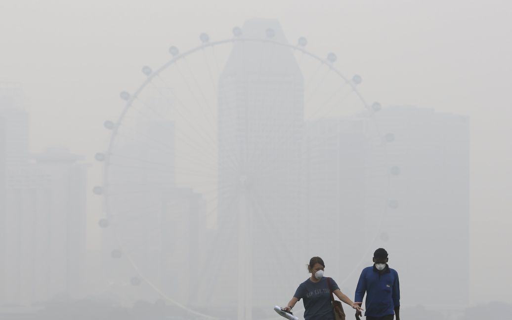 Люди в масках идут на фоне колеса обозрения Singapore Flyer в Сингапуре. Дым от лесных пожаров в Индонезии достиг соседних Малайзии и Сингапура, из-за чего уровень загрязнения воздуха в этих странах превзошел все критические нормы в несколько раз. / © Reuters