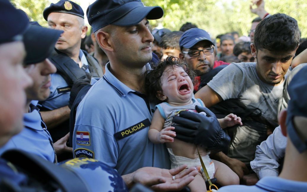 Хорватский полицейский держит ребенка-мигранта в толпе беженцев, которые ждут автобус в хорватский город Товарник. В четверг власти страны заявили, что больше не могут принять мигрантов, что вызвало недовольство у тысяч беженцев, которые стоят на границе со страной. / © Reuters